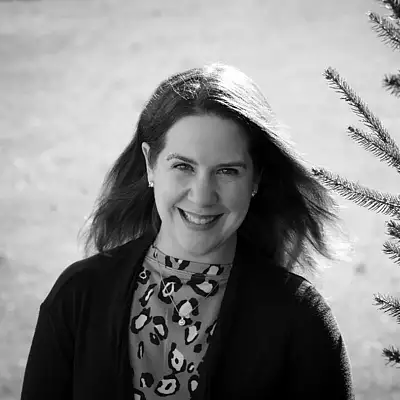 Woman smiling outdoors near a small tree, wearing a patterned top and dark cardigan. Black and white.