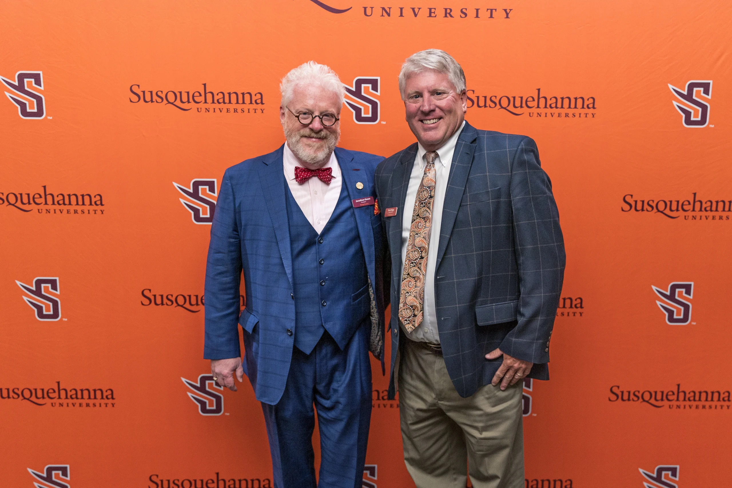 Two men in suits stand smiling in front of an orange Susquehanna University backdrop.