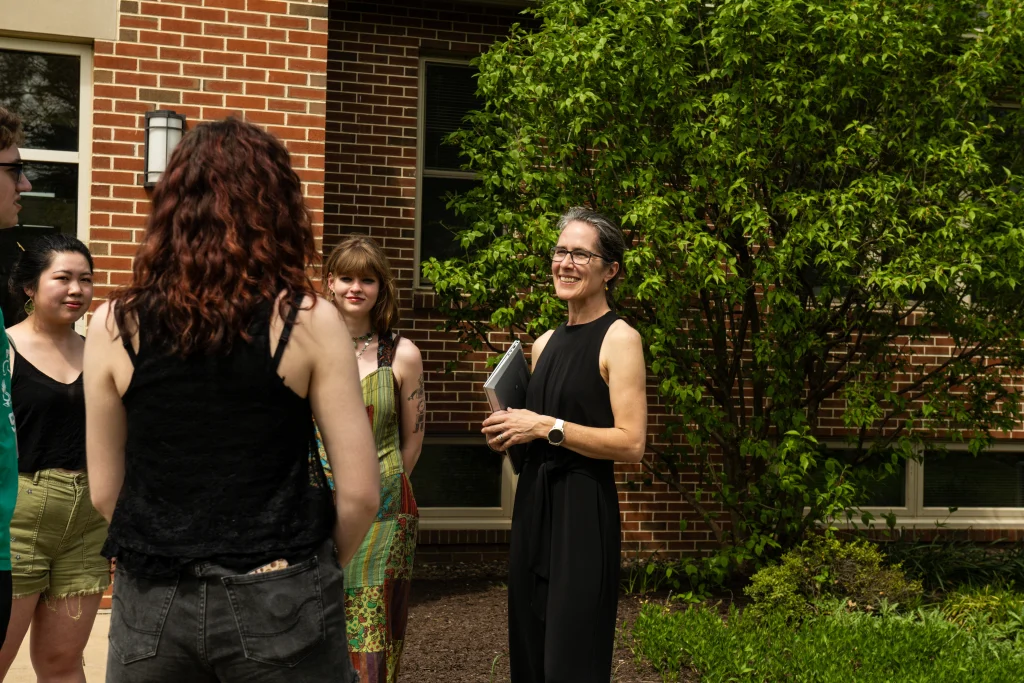 A group of people standing outside, conversing near a brick building. A person in black holds a notebook and wears glasses. Plants are visible in the background.