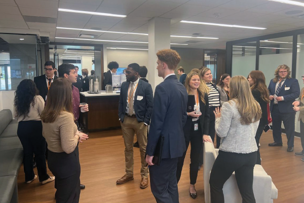 A group of people in business attire engage in conversations in an office setting with glass walls and wooden floors.