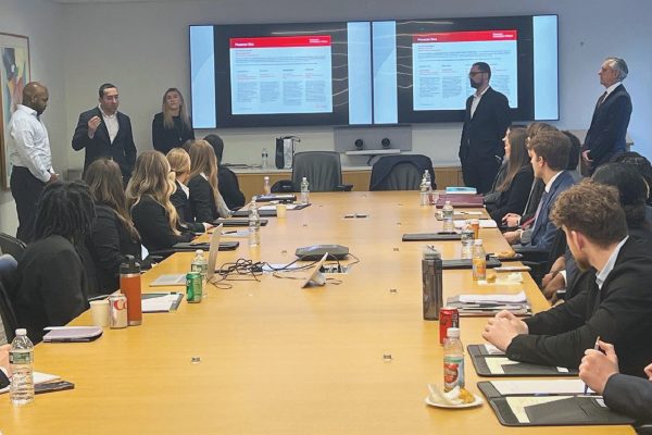 A group of people in business attire are gathered in a conference room with two large screens displaying a presentation.