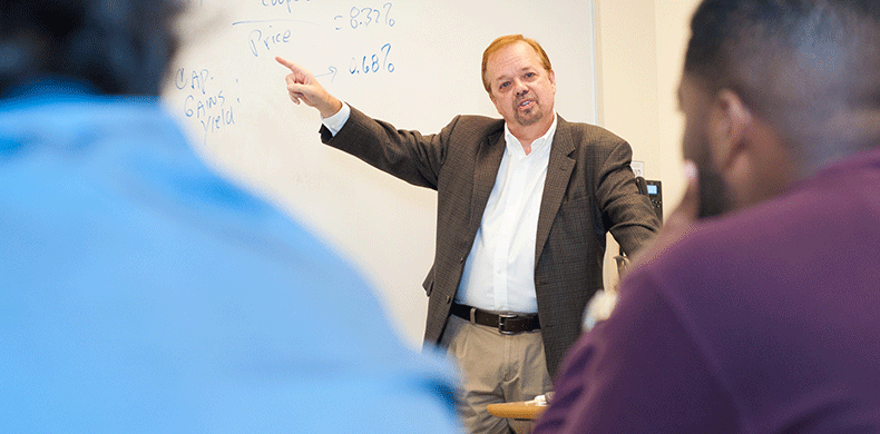 A man in a suit points to a whiteboard covered with handwritten text and numbers during a lecture. Several people sit in the foreground, listening attentively.