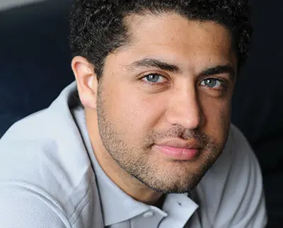 A man with curly black hair and a beard, wearing a gray collared shirt, is looking at the camera with a slight smile. The background is dark and blurred.