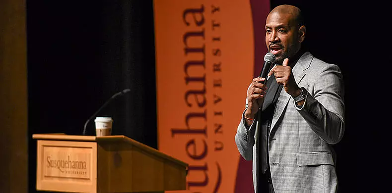 A man in a gray suit speaks into a microphone at a podium with a coffee cup, in front of a Susquehanna University banner.