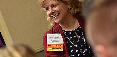 A smiling woman with curly blonde hair wears a lanyard with name tags reading "ALUMNI BOARD" and "SU Champion" at a gathering. She is dressed in a patterned top and a red cardigan.