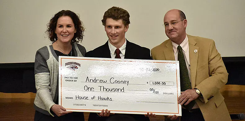 Three people stand together holding a large ceremonial check made out to Andrew Cooney for $1,000 from Susquehanna University, with “House of Hawks” written in the memo. All three are smiling at the camera.