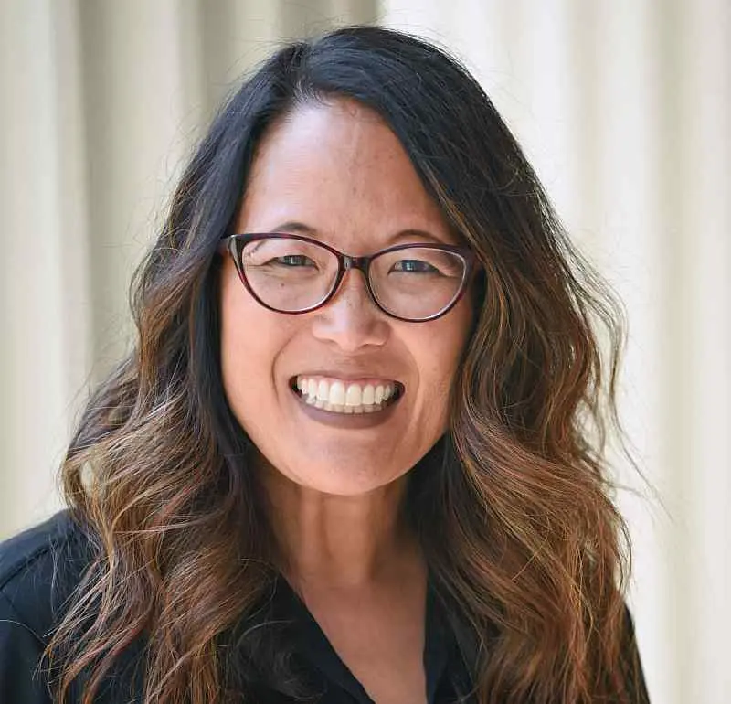 A person smiling widely, wearing glasses and a black top. They have long, wavy hair and the background is softly blurred.
