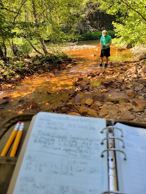 A notebook with handwritten notes and pencils is in focus in the foreground. In the background, a person wearing a green shirt stands by a rocky creek surrounded by trees under bright sunlight.