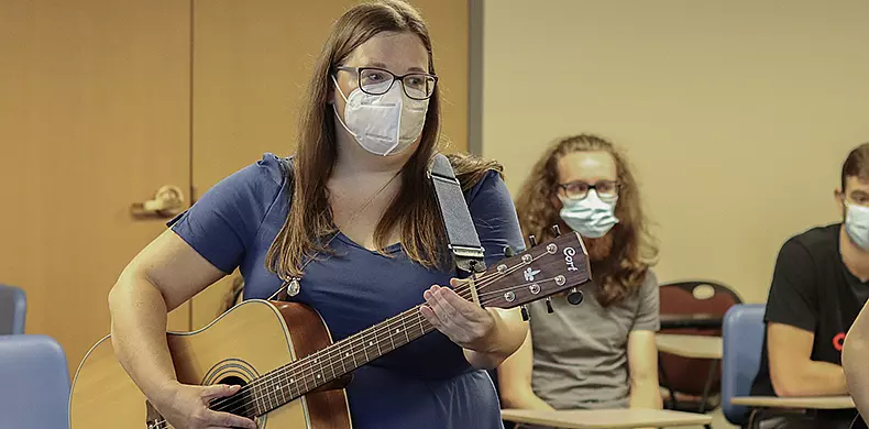 A woman wearing a mask plays an acoustic guitar in a classroom, while seated students in the background, also wearing masks, listen attentively.