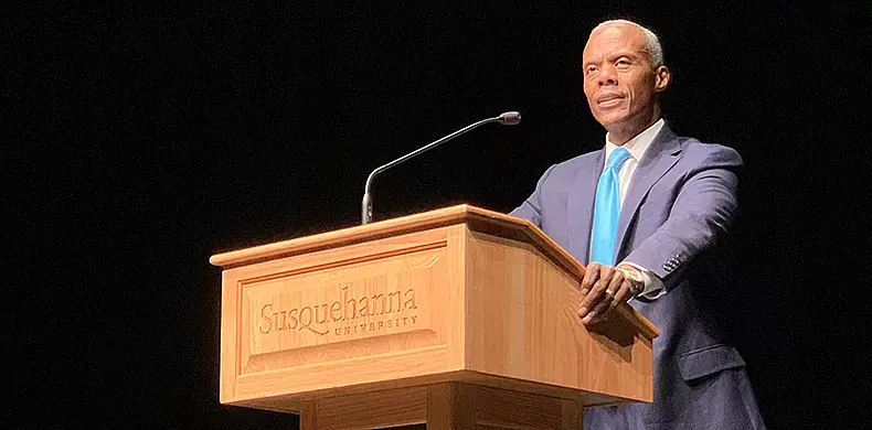 A man in a suit and blue tie stands at a wooden podium with "Susquehanna University" engraved on it, speaking into a microphone against a dark background.