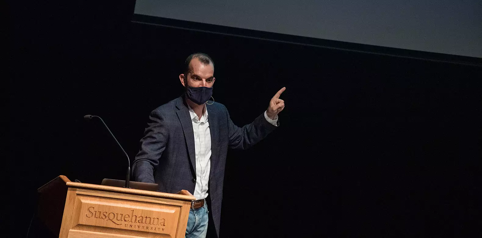 A man wearing a mask and blazer stands at a podium labeled "Susquehanna University," gesturing upward with his right hand during a presentation on a dark stage.