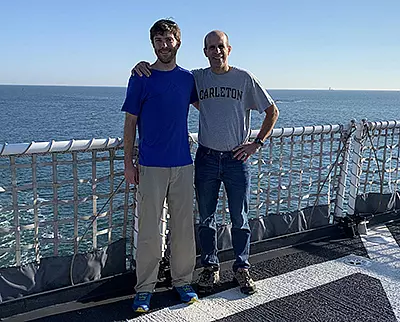 Two people stand side by side on a ship’s deck with an ocean view in the background. One person wears a blue shirt and light pants, the other a gray "Carleton" shirt and jeans. Both are smiling and appear relaxed.
