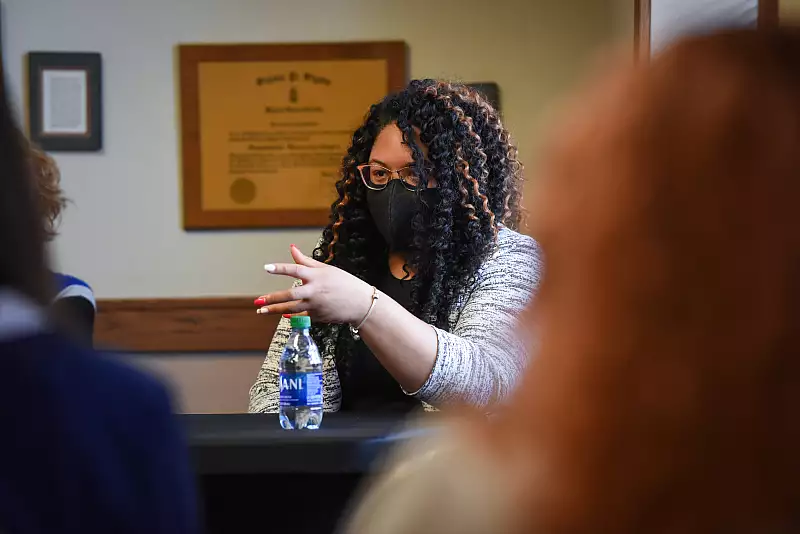 A woman with curly hair, wearing glasses and a black face mask, gestures with her hand while sitting at a table with a water bottle. Others are partially visible in the foreground.