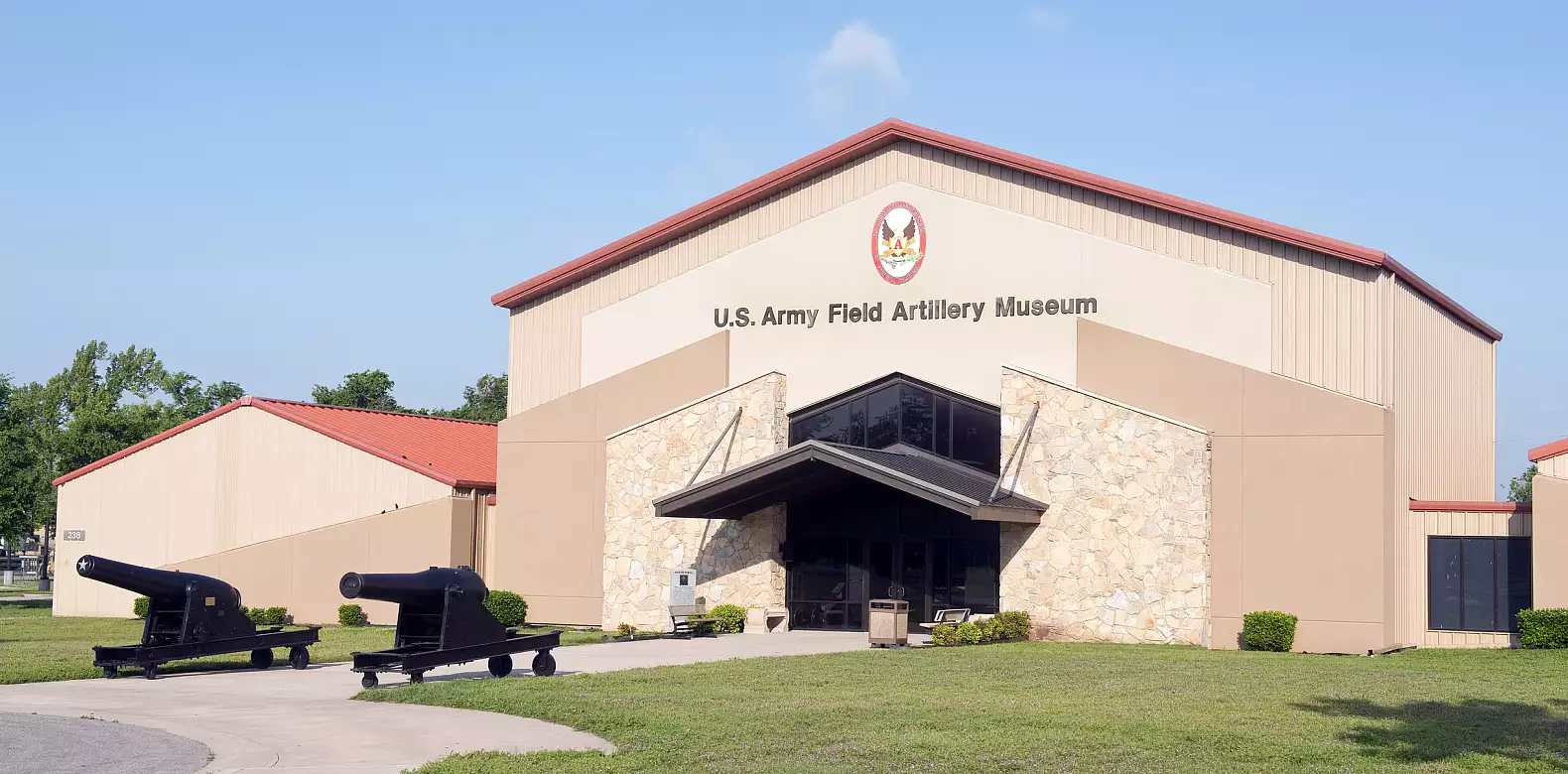 A tan building with a red roof labeled "U.S. Army Field Artillery Museum." Two black cannons are displayed on the lawn in front of the entrance. The sky is clear and blue.