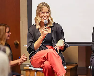 A woman sitting on a stool, holding a microphone and smiling. She is wearing a black shirt and red pants, with a relaxed posture. Another person is visible clapping in the background.