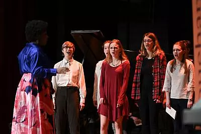 A group of five people, four women and one man, sing on stage under the direction of a person in a blue blouse and floral skirt. A piano is visible in the background, and the scene is dimly lit by stage lights.