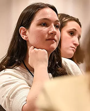 Two people are seated, attentively listening. The person in the foreground has long dark hair, rests their chin on their hand, and wears a light-colored top. The person in the background has long hair and a focused expression.