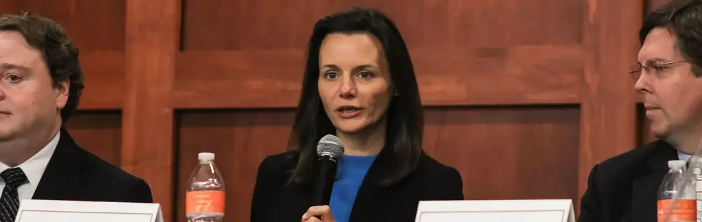 A woman holding a microphone speaks at a panel discussion. She is seated between two men, each wearing suits. The setting appears formal, with a wooden backdrop and nameplates in front of each person.