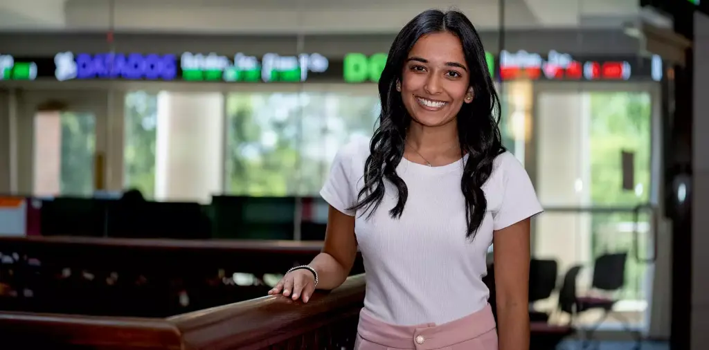 Smiling person with long black hair wearing a white shirt and pink pants stands indoors near a railing. Behind them is a stock ticker display showing stock symbols and prices.