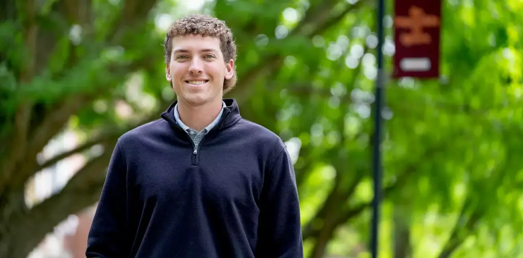 Smiling individual wearing a black sweater stands outdoors with blurred green trees and a red banner in the background.