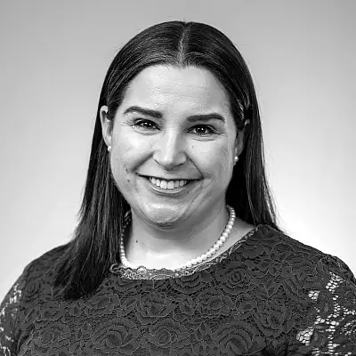 A person with long, dark hair is smiling at the camera. They are wearing a lace-patterned top and a pearl necklace. The background is plain and light-colored. The photo is in black and white.