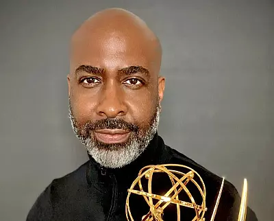 A person with a beard holds a gold Emmy award against a dark background, wearing a dark top.