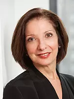 A woman with short brown hair is smiling at the camera. She is wearing a black blazer and is in a well-lit indoor setting.