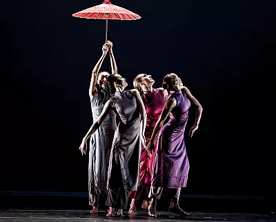 Four dancers in flowing dresses perform gracefully on stage under a red umbrella. The scene is set against a dark background, highlighting their elegant movements and colorful attire.