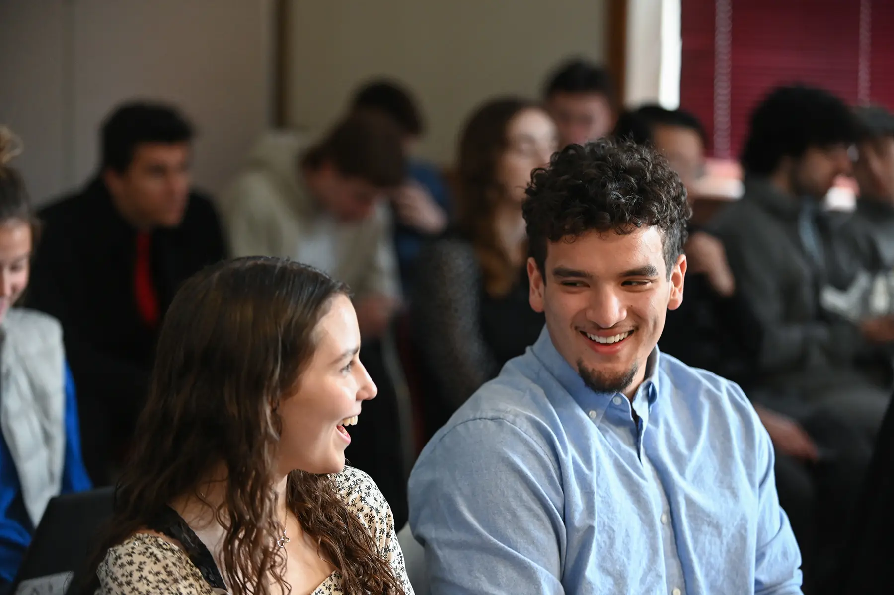 A young man and woman are smiling and talking to each other in a classroom setting. They are surrounded by other students who are seated and engaged in different activities.