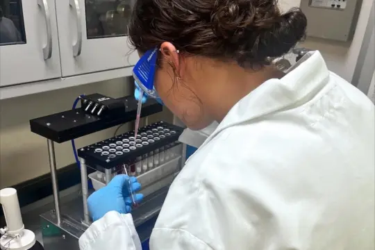 A person in a lab coat and safety goggles works with a pipette in a laboratory. They are focused on placing liquid into small vials organized in a rack. Shelves with equipment are visible in the background.