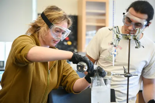 In a chemistry lab, two people don goggles for a science experiment. One carefully pours liquid from a flask into a beaker surrounded by ice, as the other observes. Both are gloved and concentrated on the intricate procedure at hand.