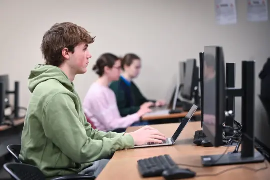 Three students sit at a row of desks with computers. The person in the foreground wears a green hoodie and works on a laptop. The other two students also focus on their screens, typing. The classroom has a plain wall with a few posters.