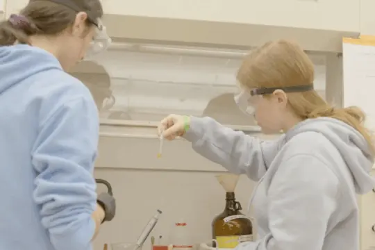 Two students wearing goggles conduct a biochemistry experiment in the laboratory. One holds a pipette, while the other observes intently. Bottles and lab equipment are scattered across the counter. Both wear casual, light-colored hoodies.