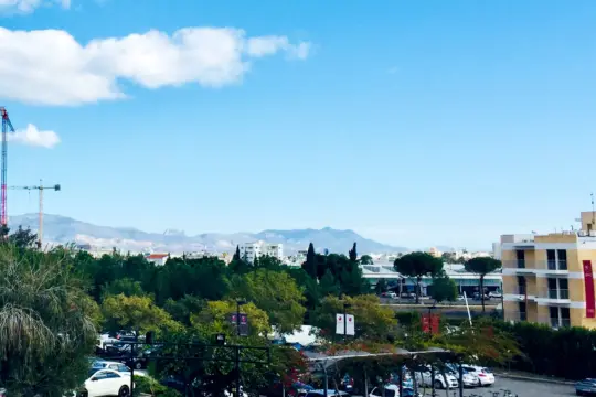 A cityscape with a clear blue sky, distant mountains, and greenery. In the foreground, parked cars and bicycles rest beneath a shelter like students in an MBA 4+1 Program, while a crane on the left stands tall among scattered buildings.