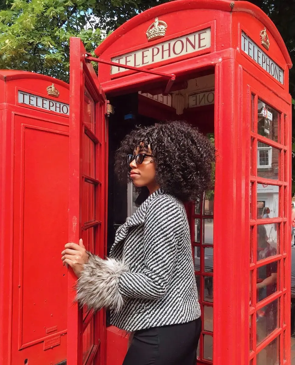 A person with curly hair and sunglasses stands in a red telephone booth, wearing a black and white textured jacket with fur accents. The iconic booth features a crown emblem and the word "TELEPHONE" above. Trees and buildings are visible in the background.