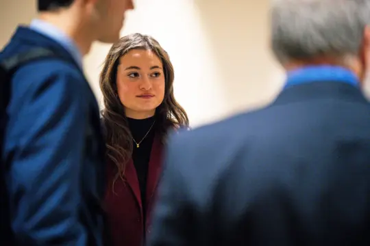 A woman with long hair wearing a maroon blazer stands among a group of people. She is looking slightly to the side with a thoughtful expression, perhaps contemplating the next step in her MBA 4+1 Program journey. The image is indoors with a neutral background.