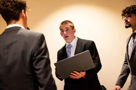 A young man in a suit holds a laptop and speaks to three other people in suits, appearing to discuss or present something. The group is standing indoors against a plain wall, engaged in conversation.