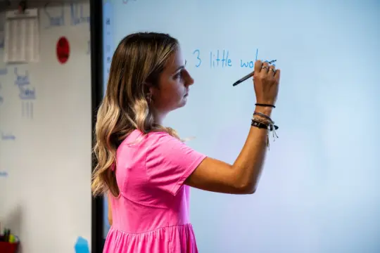 A woman in a pink dress writes on a whiteboard with a blue marker. Her hair is blonde, and she is wearing bracelets on her left wrist. The room appears to be a classroom.