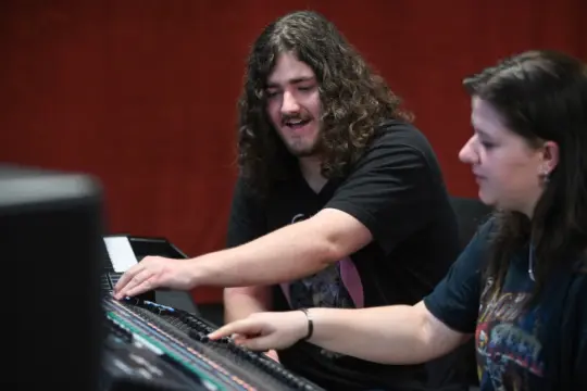 Two people are sitting at an audio mixing console. One with long curly hair is smiling and pointing at the controls, while the other person makes adjustments. They appear to be collaborating in a recording studio with red walls.