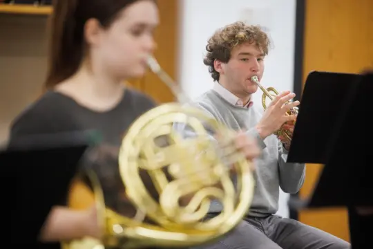 Two young musicians play brass instruments in a classroom. The focus is on a person in the background playing a trumpet, while another in the foreground plays a French horn. Music stands are visible nearby.