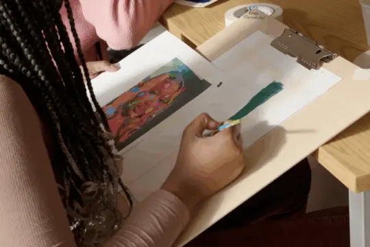 A person with braided hair is painting a green stroke on paper at a desk, inspired by a reference picture of colorful artwork reminiscent of Art History. A small palette with paint is visible on the table.