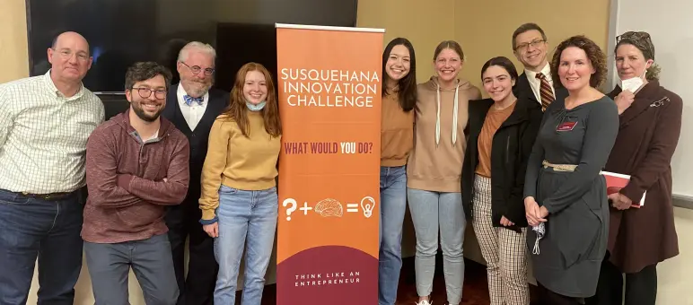 A group of people stands smiling beside a banner for the Susquehanna Innovation Challenge. The banner features a lightbulb and the phrase "What would you do?" The individuals are casually dressed, and one person holds a mask in their hand.