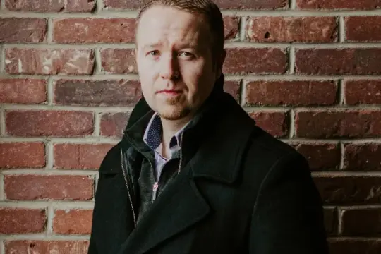 A person with short hair and a light beard stands in front of a brick wall, wearing a dark coat. They look directly at the viewer with a calm expression, and the background highlights the texture of the bricks.
