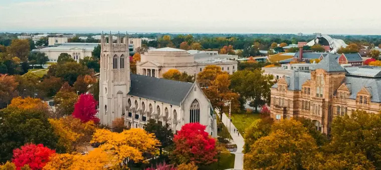 Aerial view of a university campus in autumn. Prominent gothic-style buildings are surrounded by red, yellow, and green trees. In the background, other academic buildings are visible under a cloudy sky.