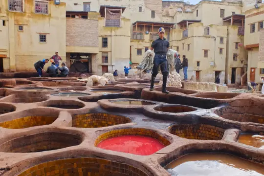 Artisan stands on the edge of a large circular vat at a traditional tannery, surrounded by other vats filled with colorful liquids and workers in the background. Old buildings with weathered walls are visible behind the tannery area.