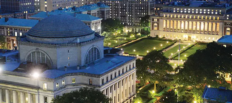 Aerial view of an illuminated university campus at night, featuring a large domed building on the left and a symmetrical, lighted courtyard with surrounding trees. Several other buildings are visible in the background.