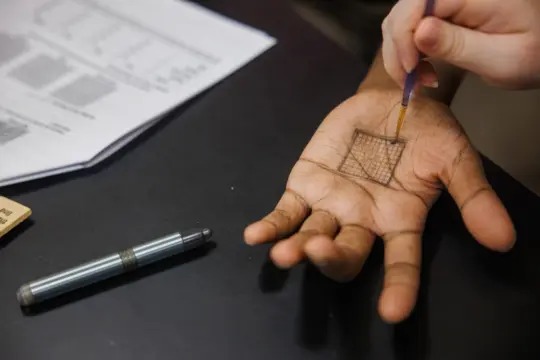 A person is drawing a grid on the palm of another person's hand using a small brush or pen. A pen and some papers with graphs are visible on the table nearby.