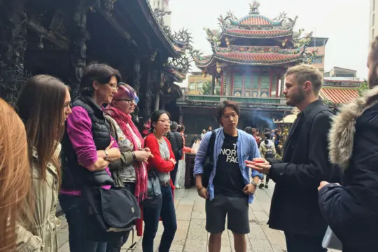 A group of people stands in conversation near a traditional temple with ornate architecture and incense burning. The scene, reminiscent of an Asian Studies discussion, appears lively and engaged against the backdrop of the cultural landmark.