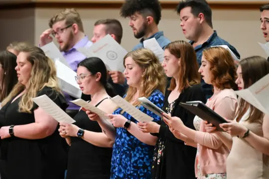 A diverse group of people sings from sheet music in a choir setting. They stand closely together, focused on their music, each holding a booklet. The background is simple, putting emphasis on the singers.