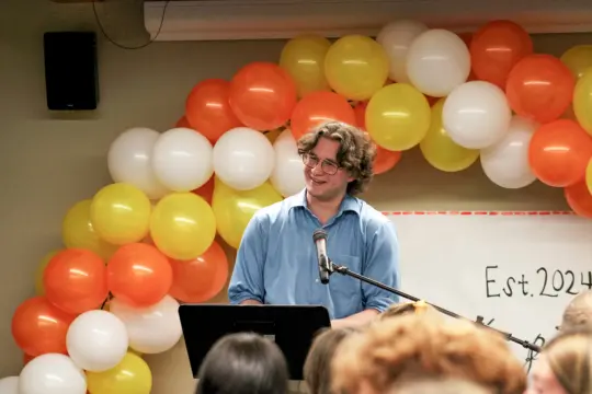 A person in a blue shirt, immersed in the art of creative writing, speaks at a podium with a microphone, smiling. Behind them, a vibrant balloon arch of orange, yellow, and white bursts with color. In the background, "Est. 2024" is inscribed on a whiteboard as eager audience members listen intently.